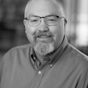 Photo of a white man with glasses, greying mustache and goatee, and a shaved head, wearing a blue button-down shirt.