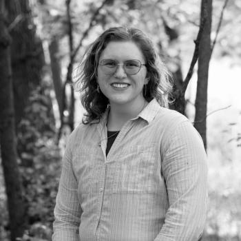 Micah Land, a white person with shoulder length curly hair, is smiling in front of sunlit woods. They are wearing a light blue button up shirt and glasses.